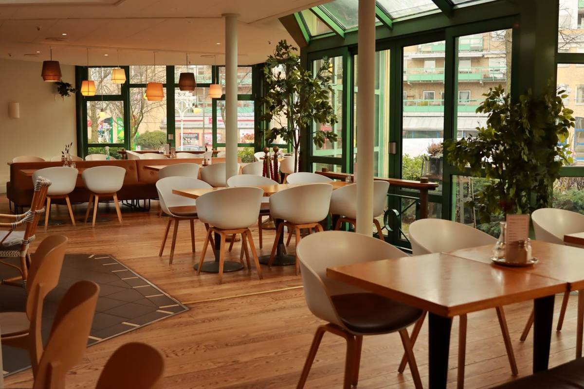 Åbent spiseområde med træborde, lyse stole og grønne planter ved glaspartier