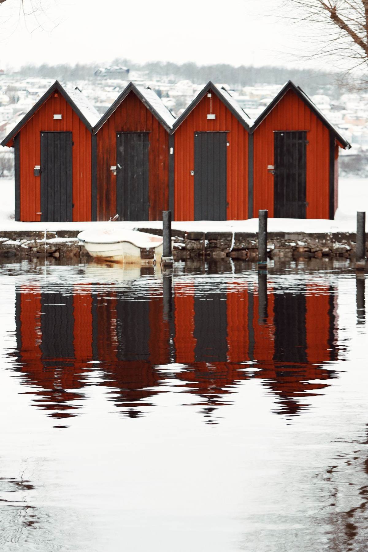 Fire røde bådhuse i sne med spejling i vandet