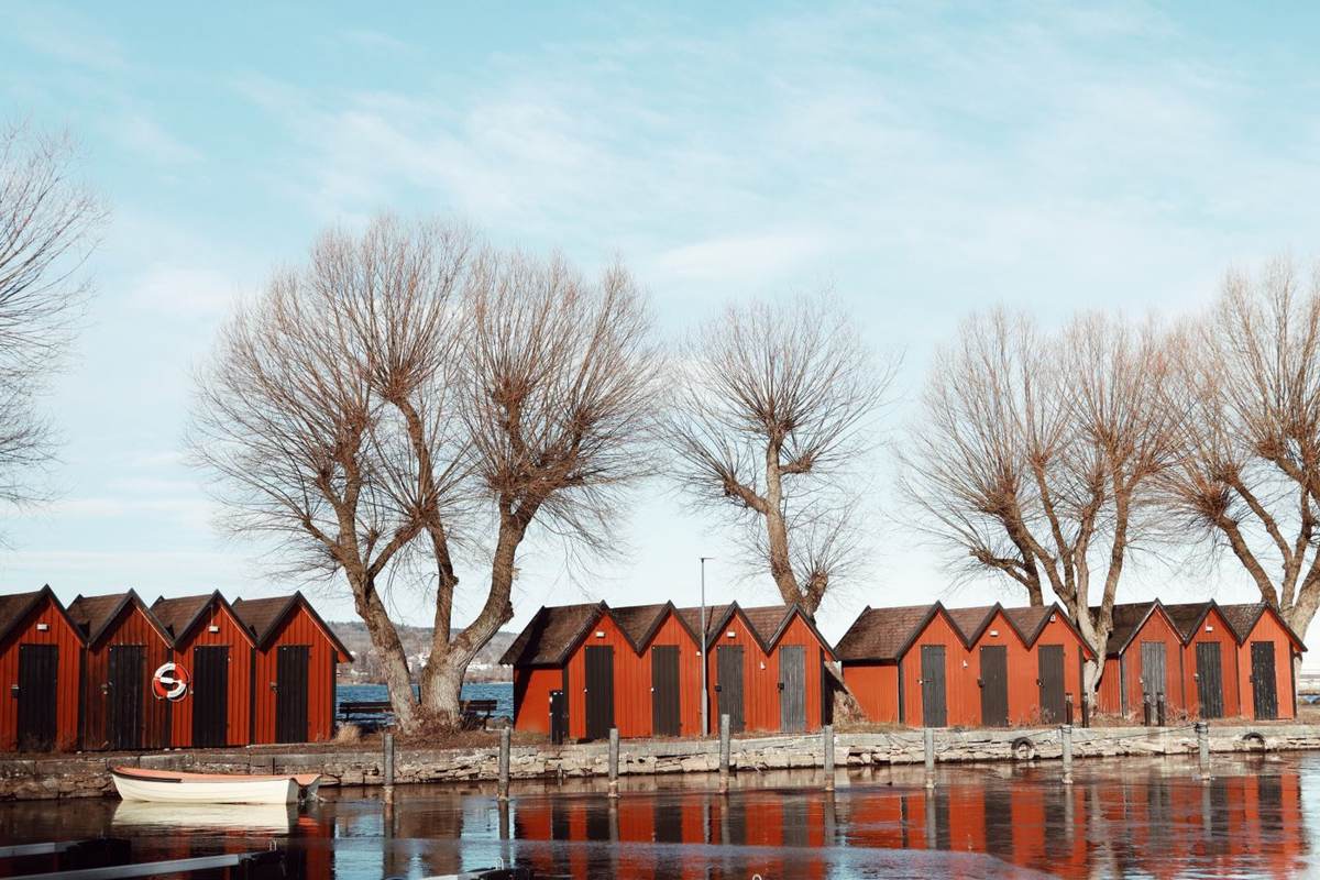 Røde bådhuse langs en sø med bare træer og blå himmel