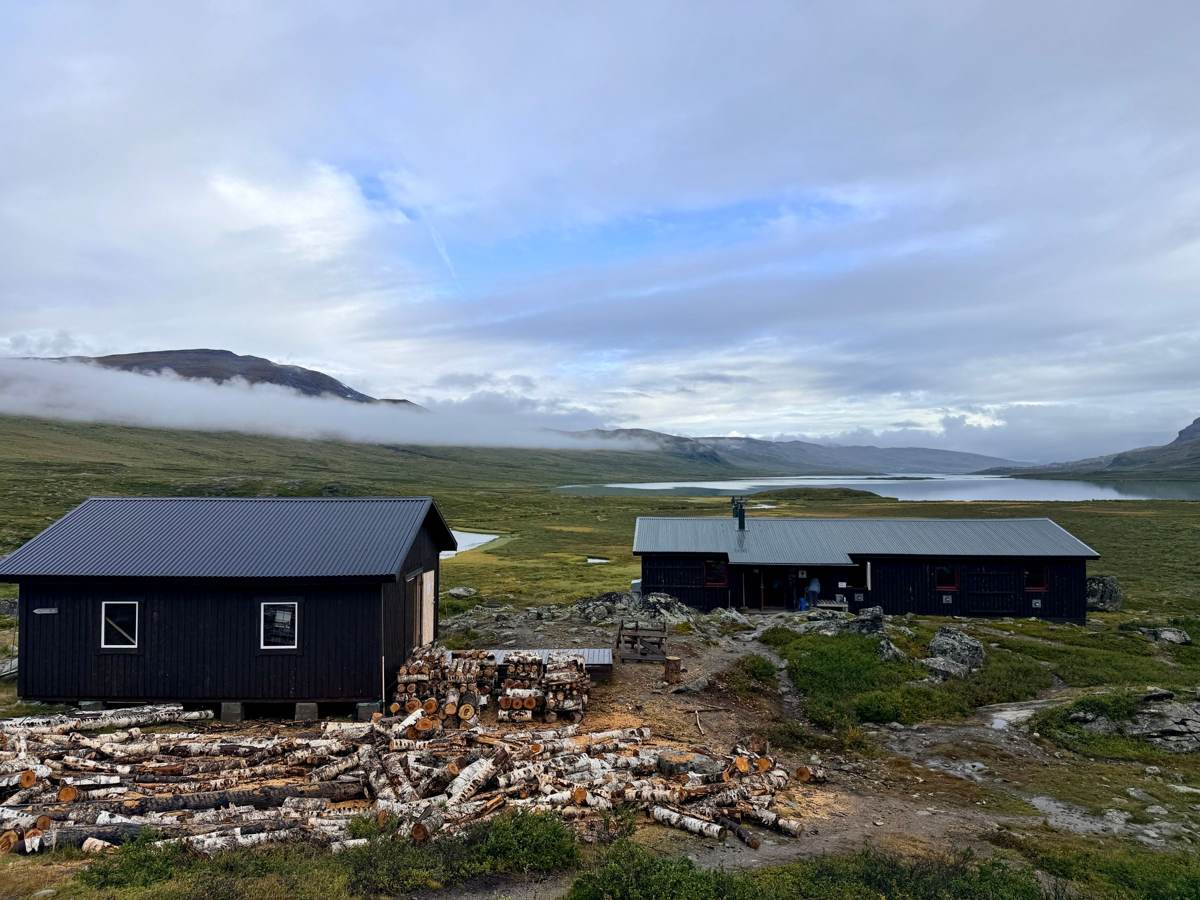 To mørke hytter i fjeldet under en skyet himmel med brænde i forgrunden