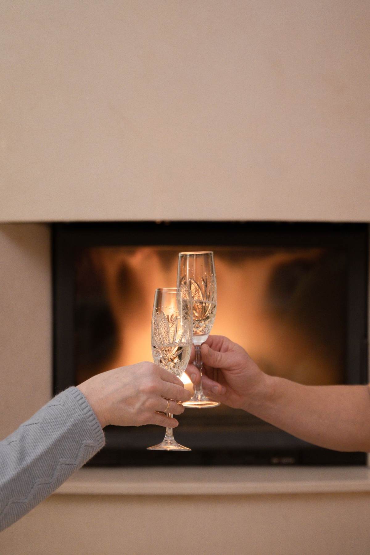 To hænder rækker champagneglas frem foran en brændeovn