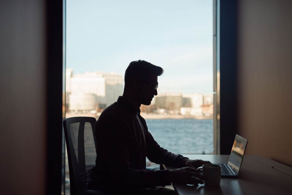 En mand sidder i silhuet ved et vindue med udsigt over vandet og arbejder på sin bærbare