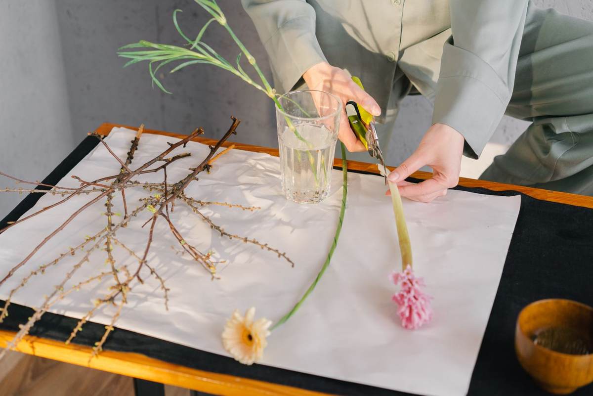 To hænder sætter blomster og kviste i en glasvase på hvidt papir