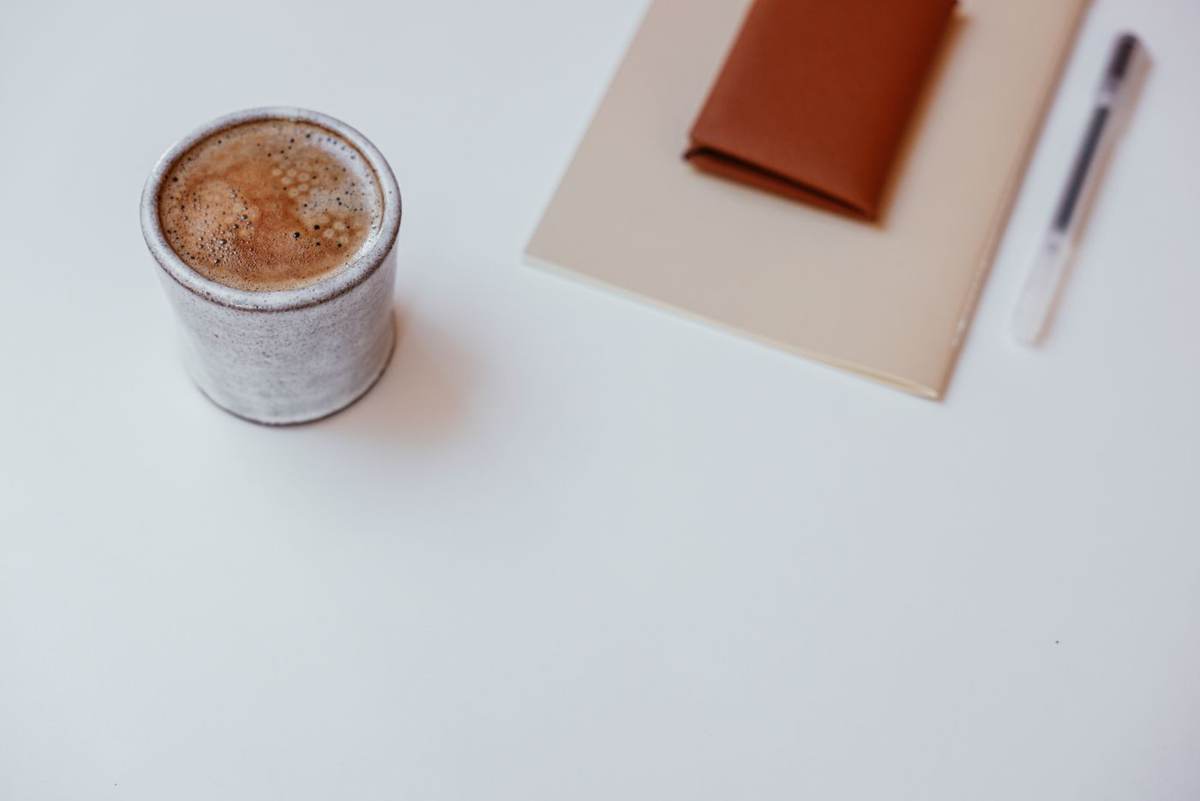 En kop kaffe, en notesbog og en pen set oppefra på et hvidt bord