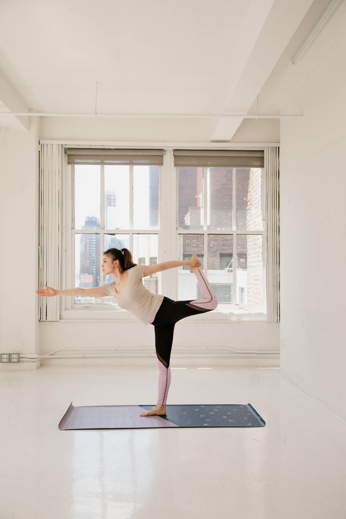 En kvinde står på ét ben på en yogamåtte i et stort, lyst rum med vinduer