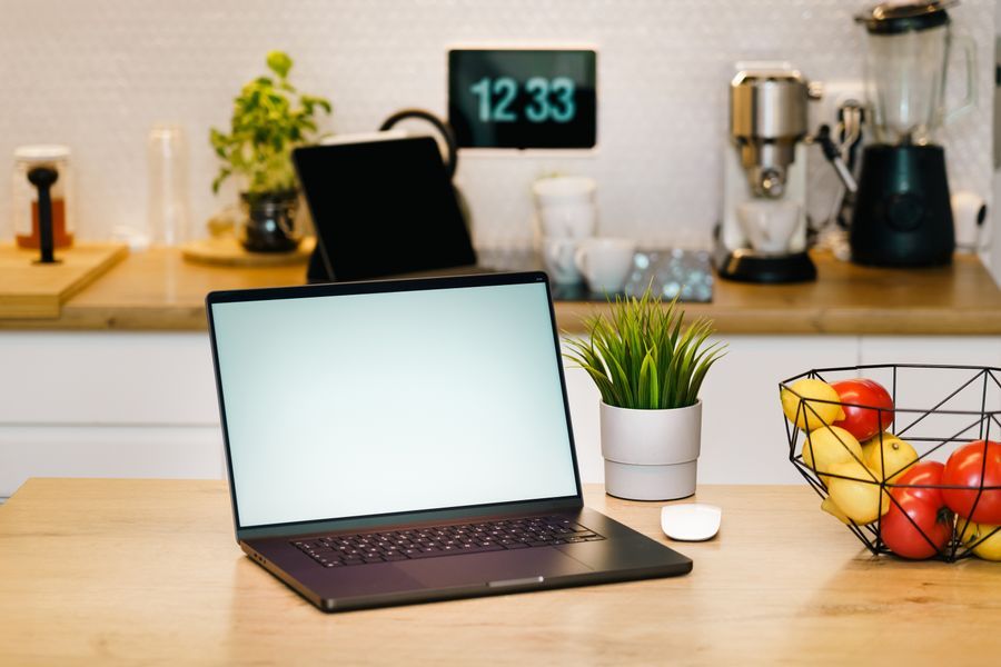 En bærbar computer står på et køkkenbord ved siden af en kaffemaskine og en skål med frugt