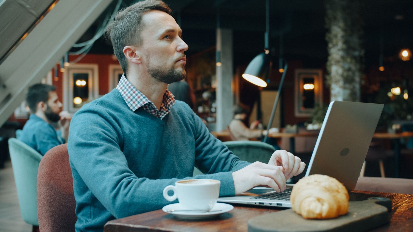 En mand arbejder på en bærbar computer ved et bord i en hyggelig café med en kop kaffe og en croissant
