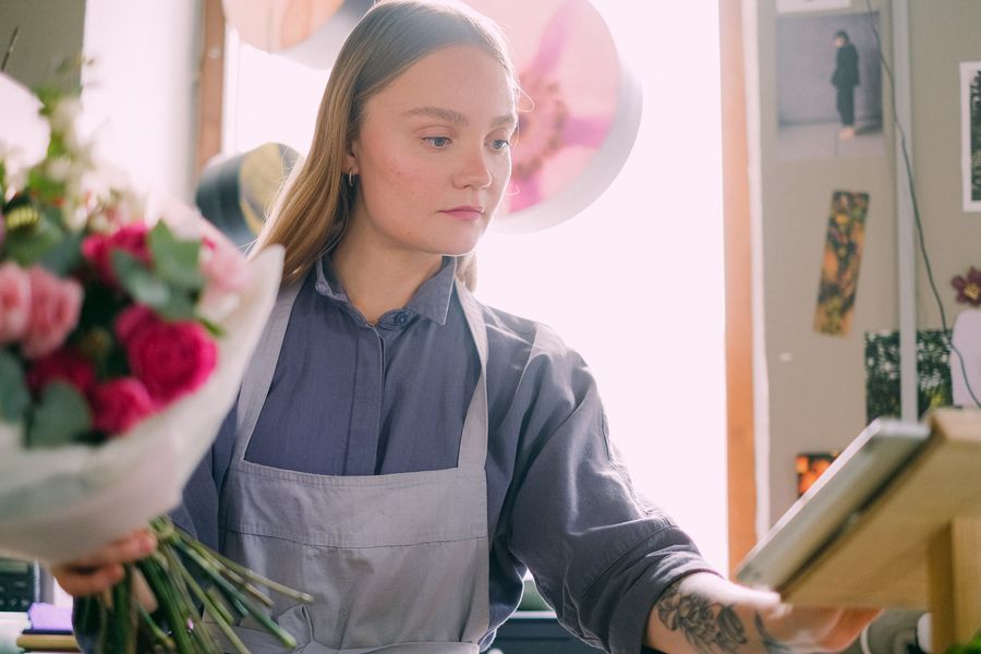 En blomsterhandler i forklæde pakker en buket ind og kigger på en tablet i en lille butik