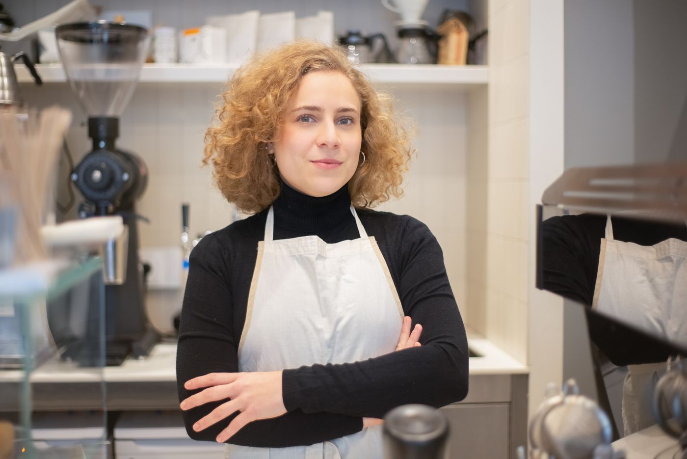 En kvinde i forklæde står med korslagte arme foran en kaffemaskine i en lille café